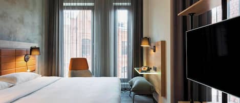 A hotel room with a large bed, a desk with a lamp, a TV, and a view of the city through large windows.