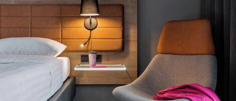 Une chambre d’hôtel avec un lit, une table de chevet avec une lampe, une chaise et une petite table sur laquelle se trouvent une tasse et un objet rose.