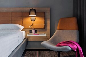 A hotel room with a bed, a nightstand with a lamp, a chair, and a small table with a cup and a pink item on it.