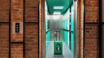 An elevator with a green suitcase and a "Good Vibes Only" sticker, surrounded by brick walls and metal mesh panels.