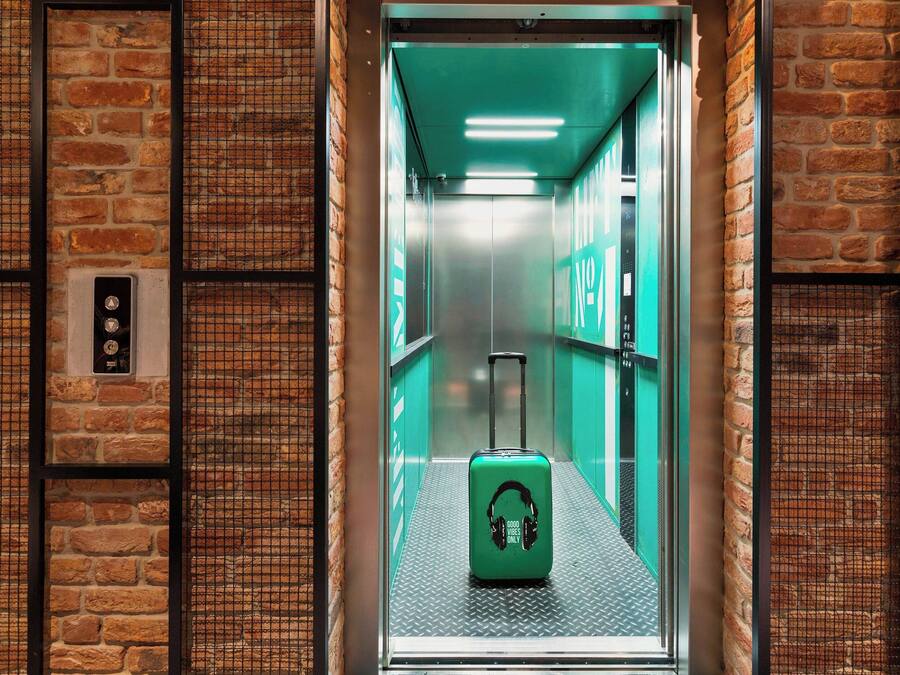 An elevator with a green suitcase and a "Good Vibes Only" sticker, surrounded by brick walls and metal mesh panels.