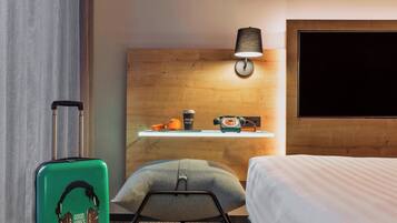 Una habitación de hotel con una cama, un escritorio con lámpara, una maleta y un almohadón.