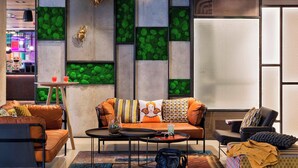 A modern living room with a sofa, coffee table, and chairs. There is a small table with a drink and a decorative item. The room has a unique green wall design and a large window.