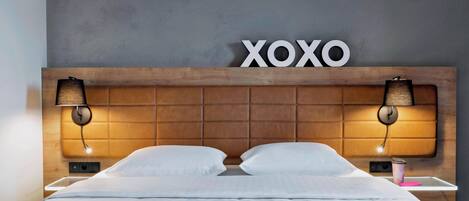 Et hotelværelse med sengegavl i træ, to senelamper, en seng med hvide sengetøj og et ophængt skilt med teksten "XOXO".