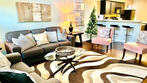 A living room with a sofa, armchairs, a coffee table, and a rug. There is a small table with a lamp, a Christmas tree, and a kitchen area in the background.