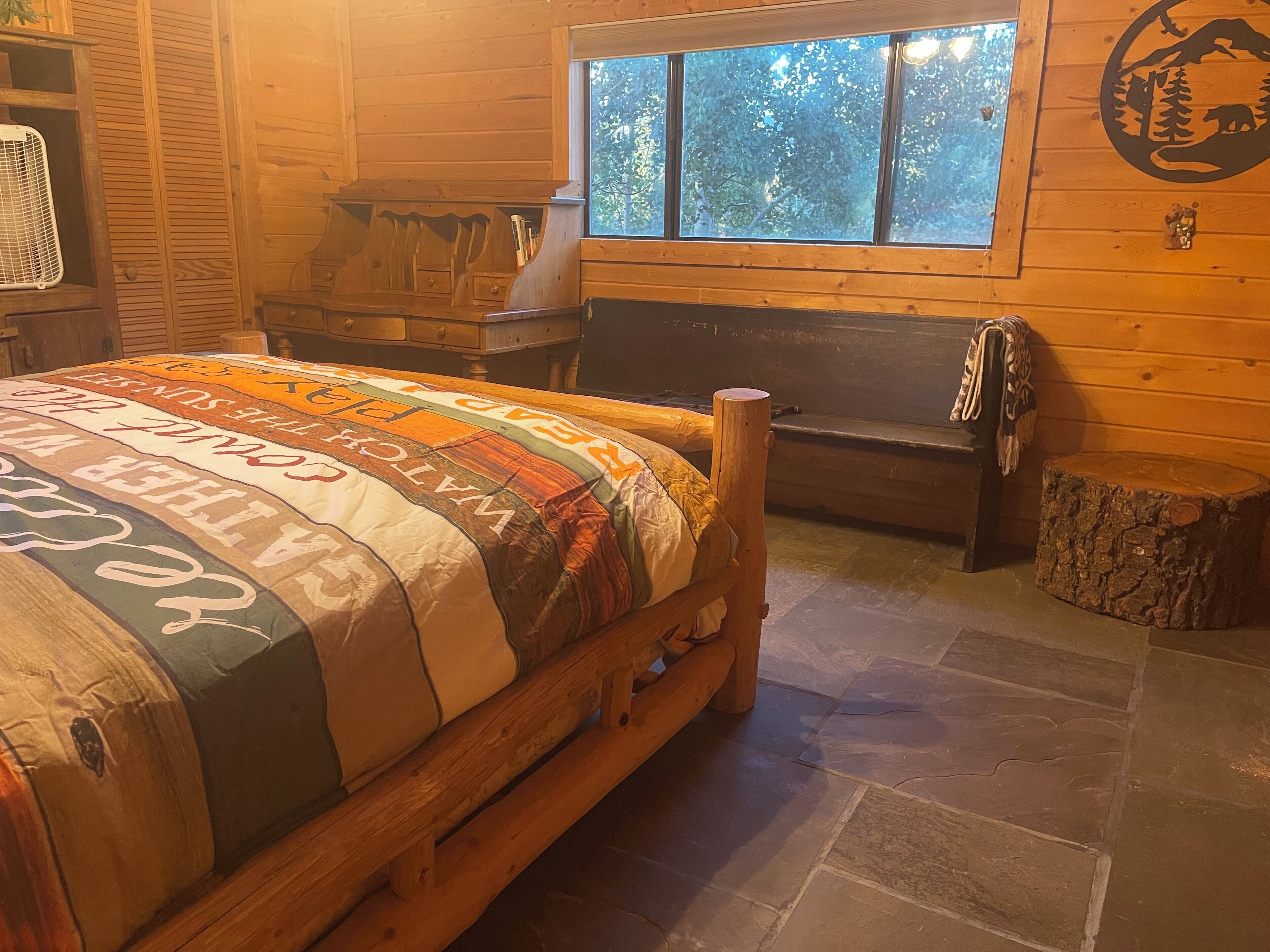 A cozy log cabin room with a wooden bed, a stone fireplace, a window with a view of trees, and a rustic wall decor.
