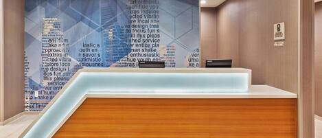A reception desk with a wooden finish and a glass ledge, set against a wall with a geometric pattern and text.