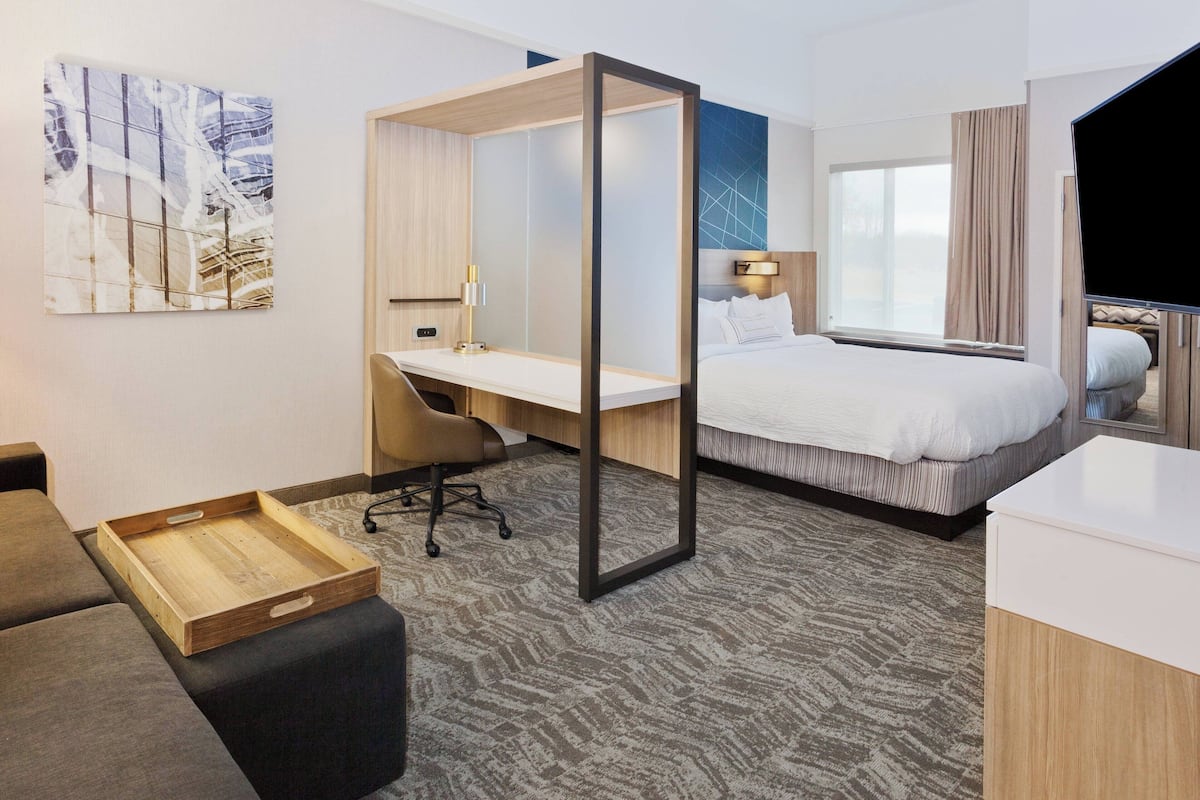 A hotel room with a bed, a desk, a chair, a wooden side table, and a large window.