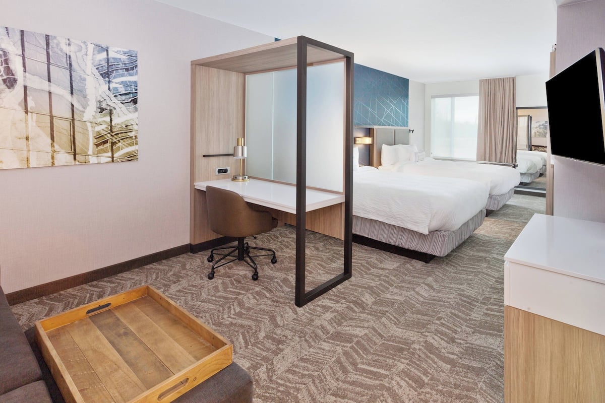A hotel room with a desk, two beds, a TV, and a large window.