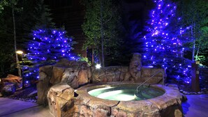 Un bain à remous installé sur un terrain rocheux, avec des arbres illuminés en arrière-plan, le tout entouré d’un mur de pierres.