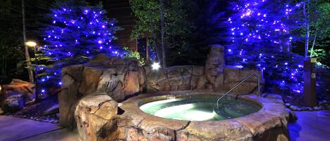 Un jacuzzi con un paisaje rocoso al fondo, árboles iluminados y un muro de piedra alrededor.