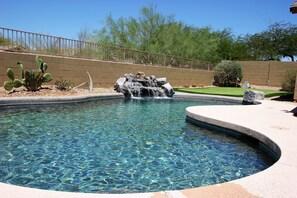 A clear swimming pool with a waterfall feature, surrounded by a brick wall and a fenced area with cacti and trees. - Beautiful Estrella Mountain Home with Private Pool and Putting Green (Goodyear)