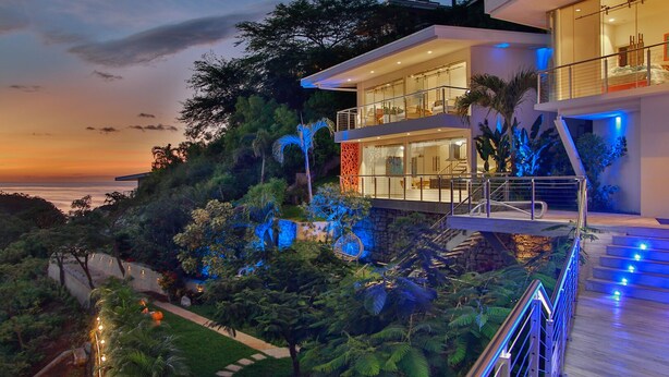 Ein modernes Haus mit einem großen Balkon, der einen üppigen Garten und einen Sonnenuntergangsansicht über das Meer bietet.