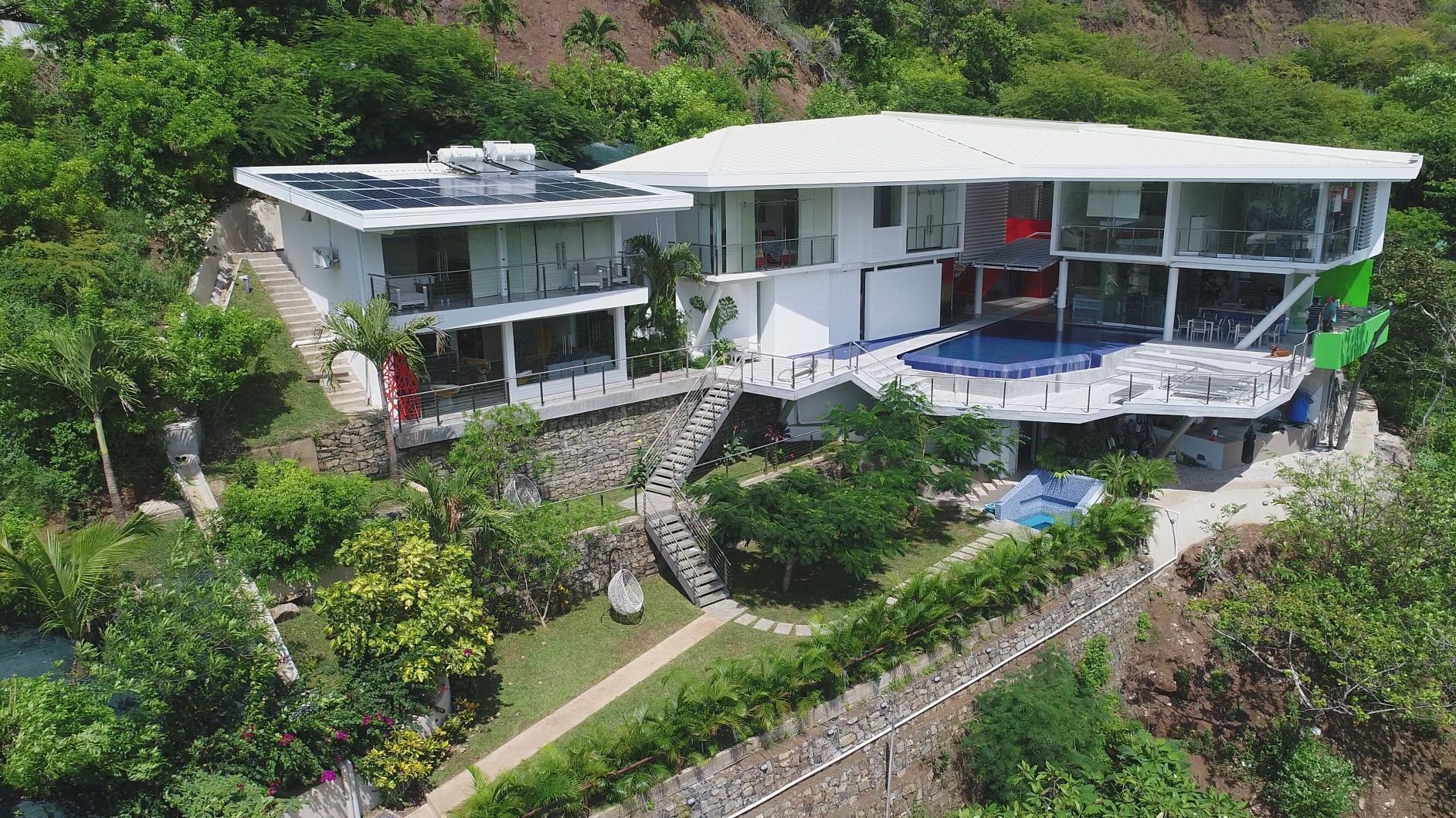 Un bâtiment moderne à deux étages, doté d’une piscine sur le toit, entouré d’une végétation luxuriante et d’un mur de soutènement en pierre.