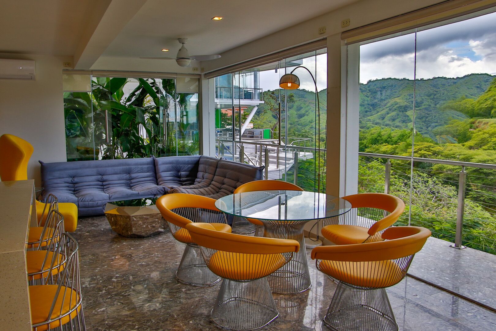 A modern living room with a glass table and orange chairs, a blue sofa, and a view of lush green hills.