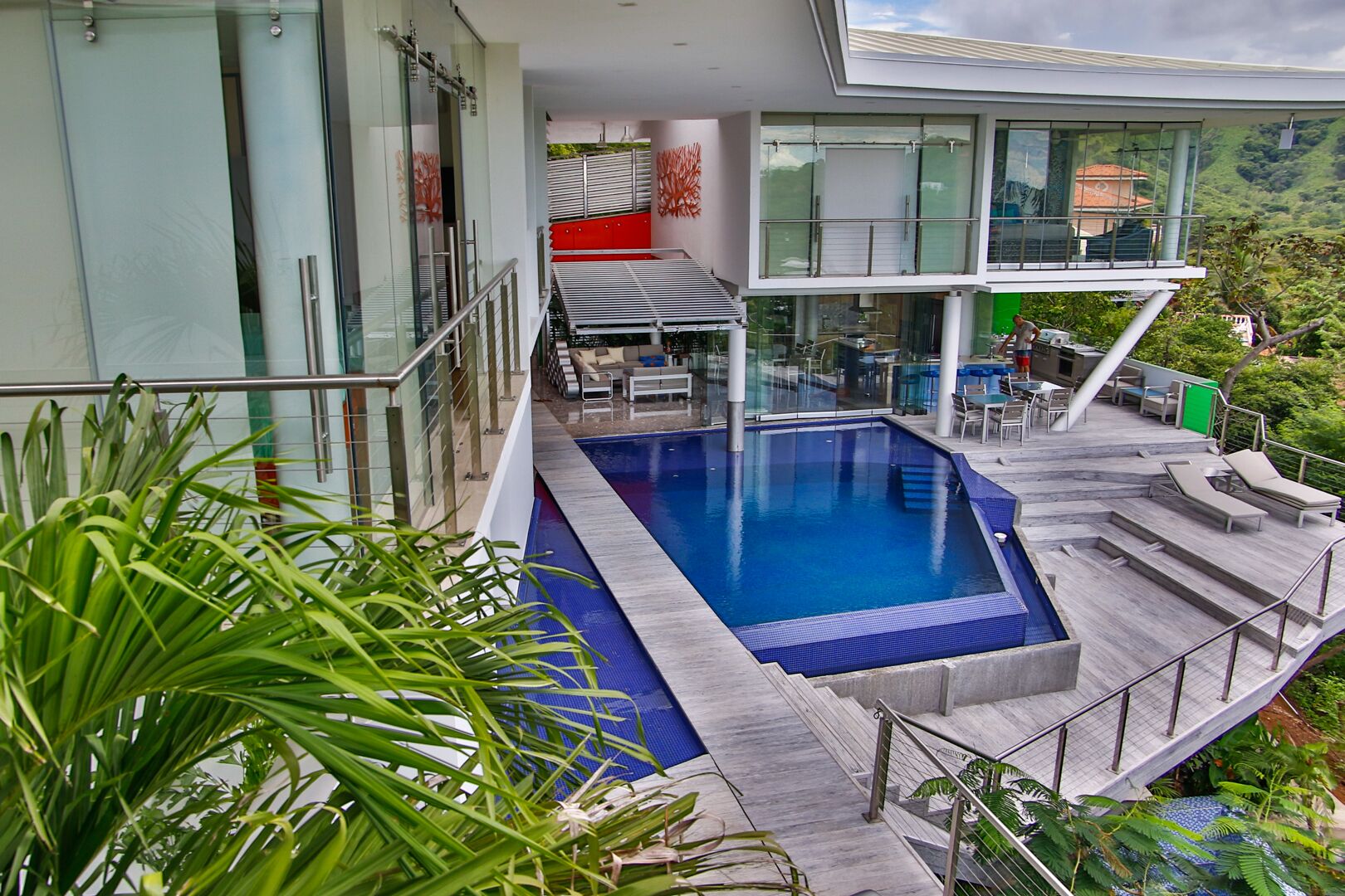 A modern outdoor pool area with a glass balustrade, lounge chairs, and a dining area with outdoor seating.