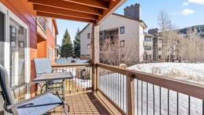 A wooden balcony with a table and chairs, overlooking a snowy landscape and neighboring buildings.