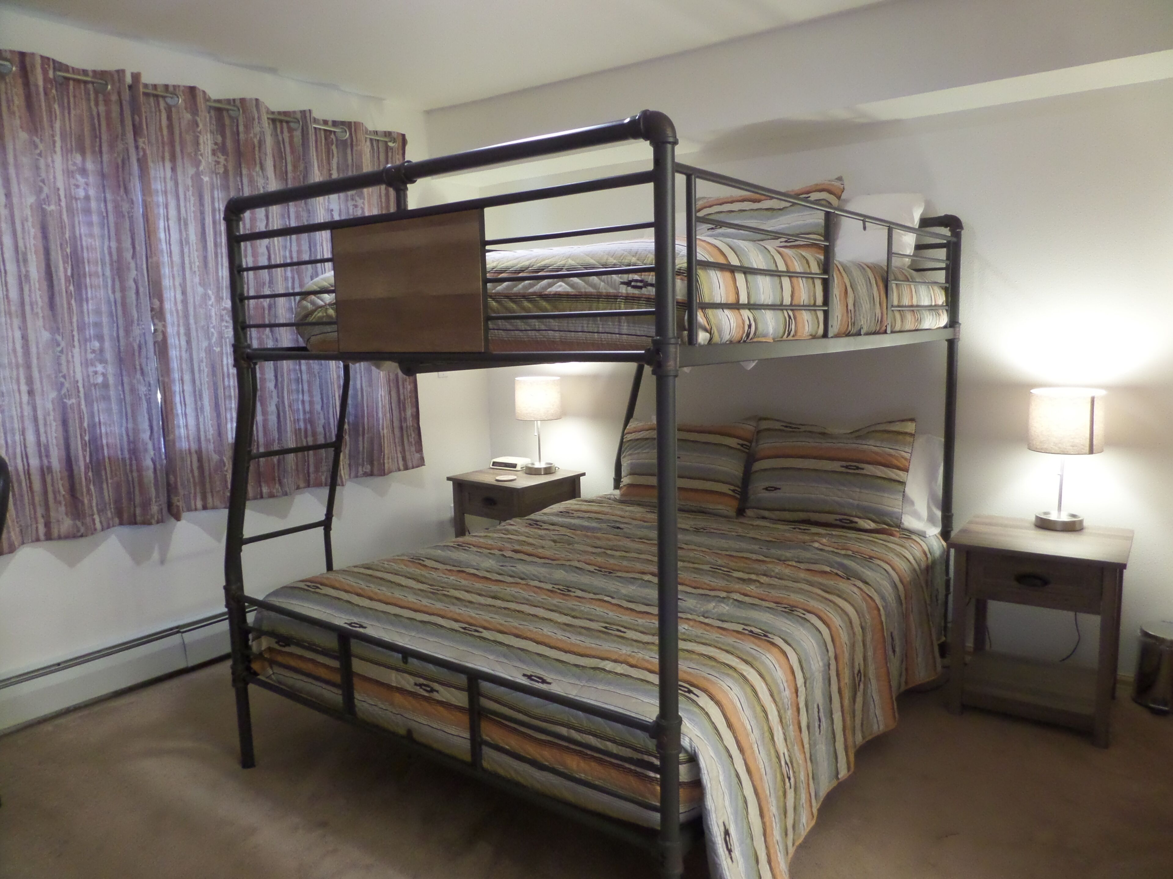 A bunk bed room with a striped bedspread, two bedside tables with lamps, and a curtain on the window.