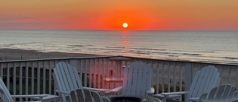 A sunset view from a deck with white Adirondack chairs and a fire pit.