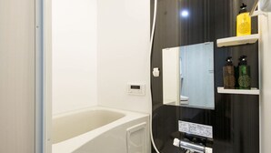 A modern bathroom with a bathtub, a mirror, and shelves with toiletries. - Trip Pod Chiyo A (Fukuoka)