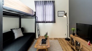 A compact living space with a bunk bed, a sofa, a coffee table with mugs, a TV on a wooden stand, and a potted plant. - Trip Pod Chiyo A (Fukuoka)