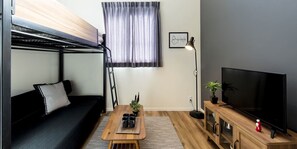 Un espace de vie compact comprenant un lit superposé, un canapé, une table basse avec des tasses, un téléviseur posé sur un meuble en bois et une plante en pot.
