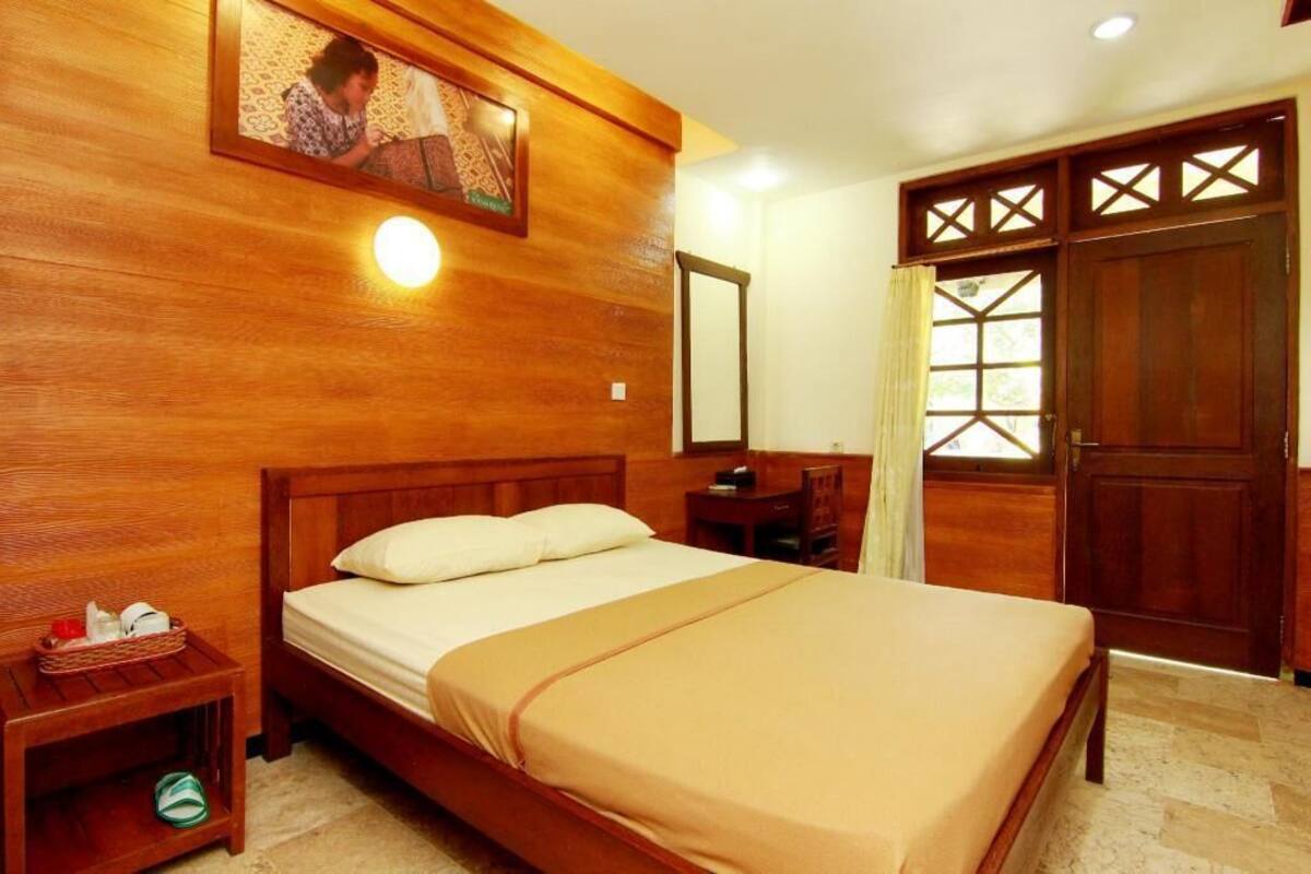 Una cama con estructura de madera y cubrecama color beige, una mesita de noche con una cesta, un espejo y una puerta de madera con un ventanal.