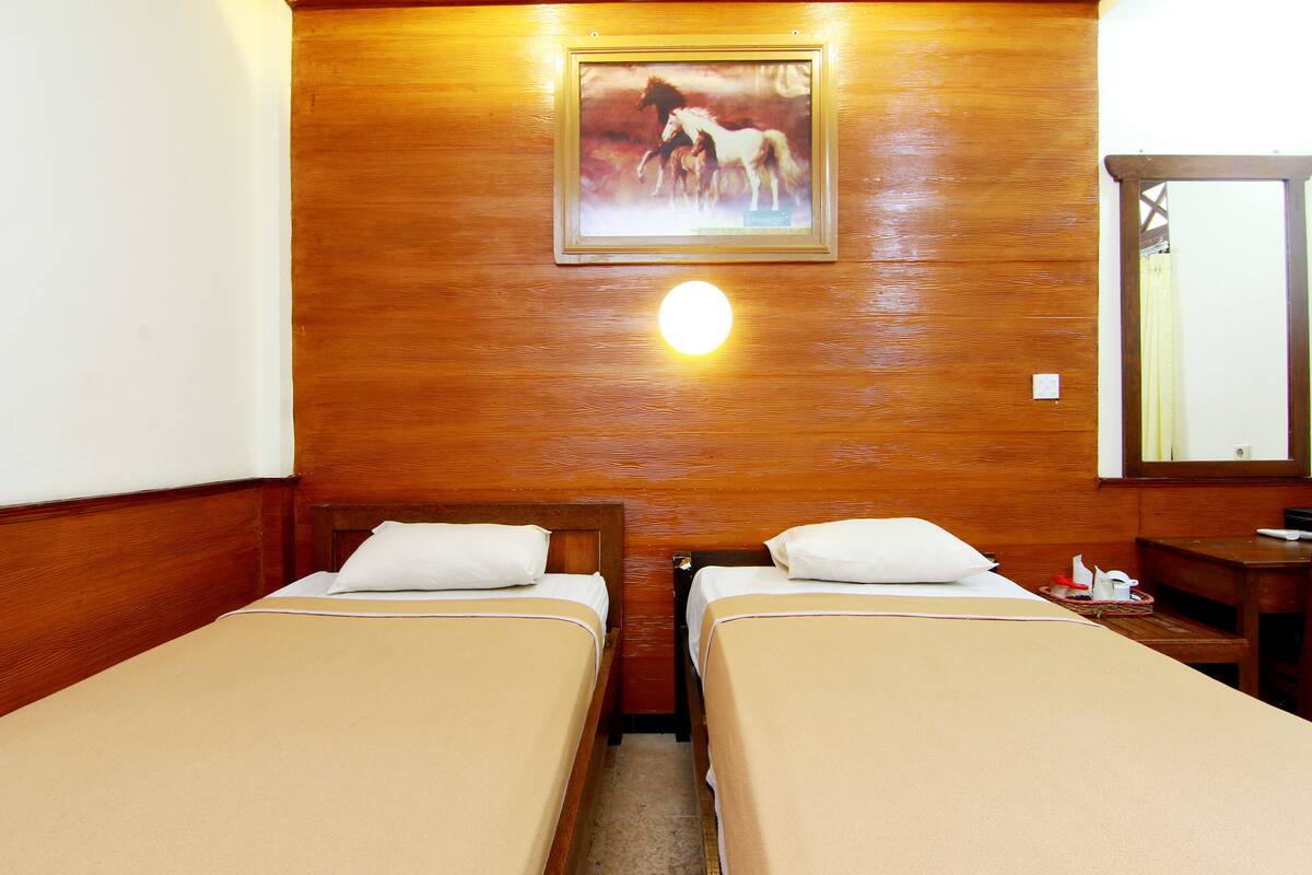 Una habitación de hotel con dos camas individuales, cabeceros de madera, una pared de madera, un cuadro enmarcado con caballos, una lámpara de pared redonda y una mesita de noche de madera con una lámpara.