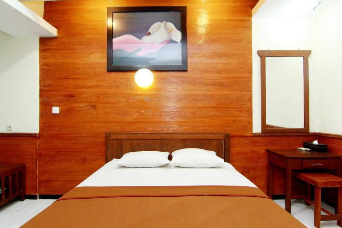 Una habitación de hotel con cabecera de madera, cama con ropa blanca, mesita de noche con lámpara y un cuadro en la pared.