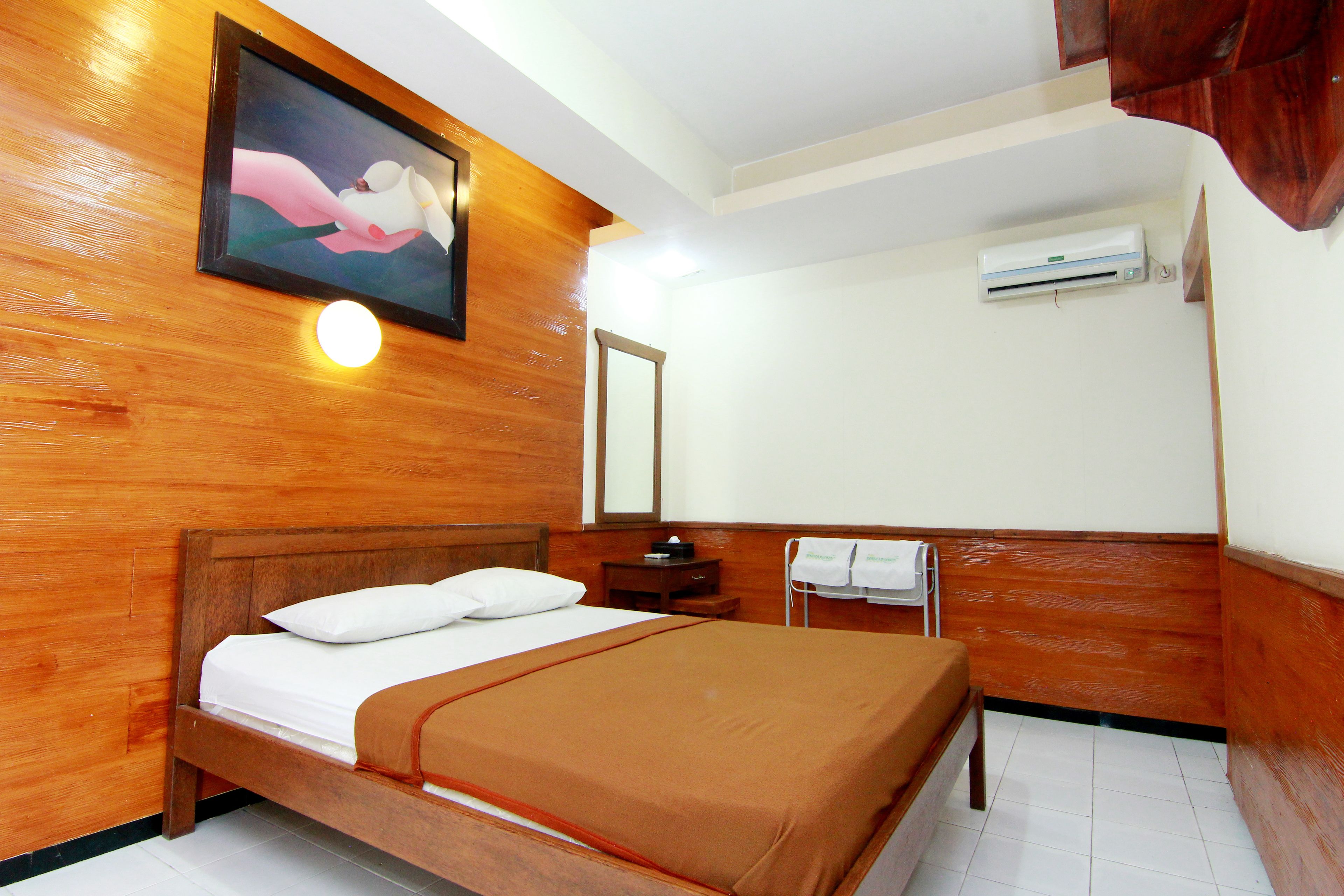 A hotel room with a wooden bed, a nightstand, a mirror, and an air conditioning unit on the wall.