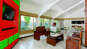 A spacious living area with a large window, a wooden bench, a coffee table, and a TV mounted on a red and green wall. - The Soemarsono Hotel (Depok)