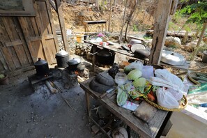 Una cocina al aire libre rústica con ollas de cocina, una mesa de madera con verduras y bolsas de plástico, y una estructura de madera al fondo.