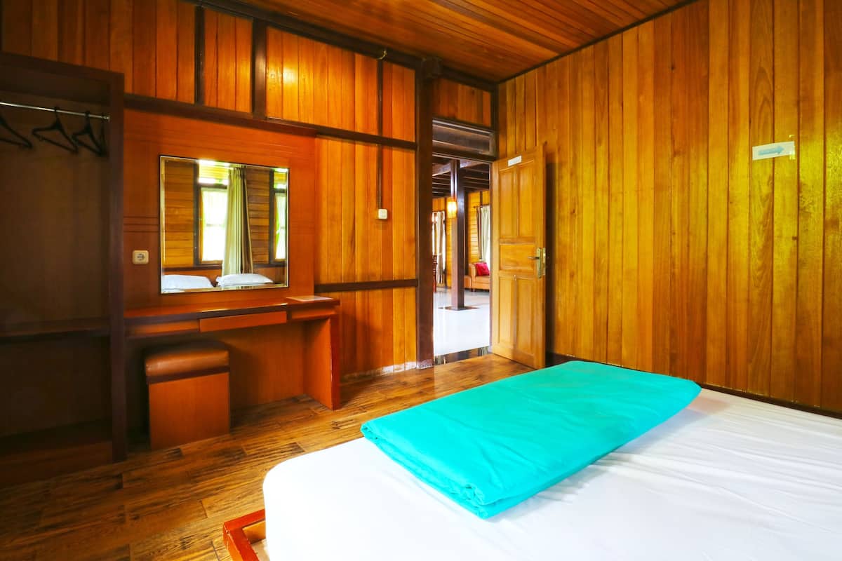 Une pièce aux murs en bois, un lit recouvert d’une couverture bleue, un bureau avec un miroir et une porte qui donne sur une autre pièce.