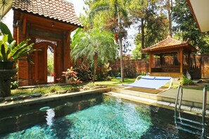 Une piscine aux eaux cristallines, dotée d’une terrasse en bois et de chaises longues, entourée d’une végétation luxuriante et de constructions traditionnelles en bois.