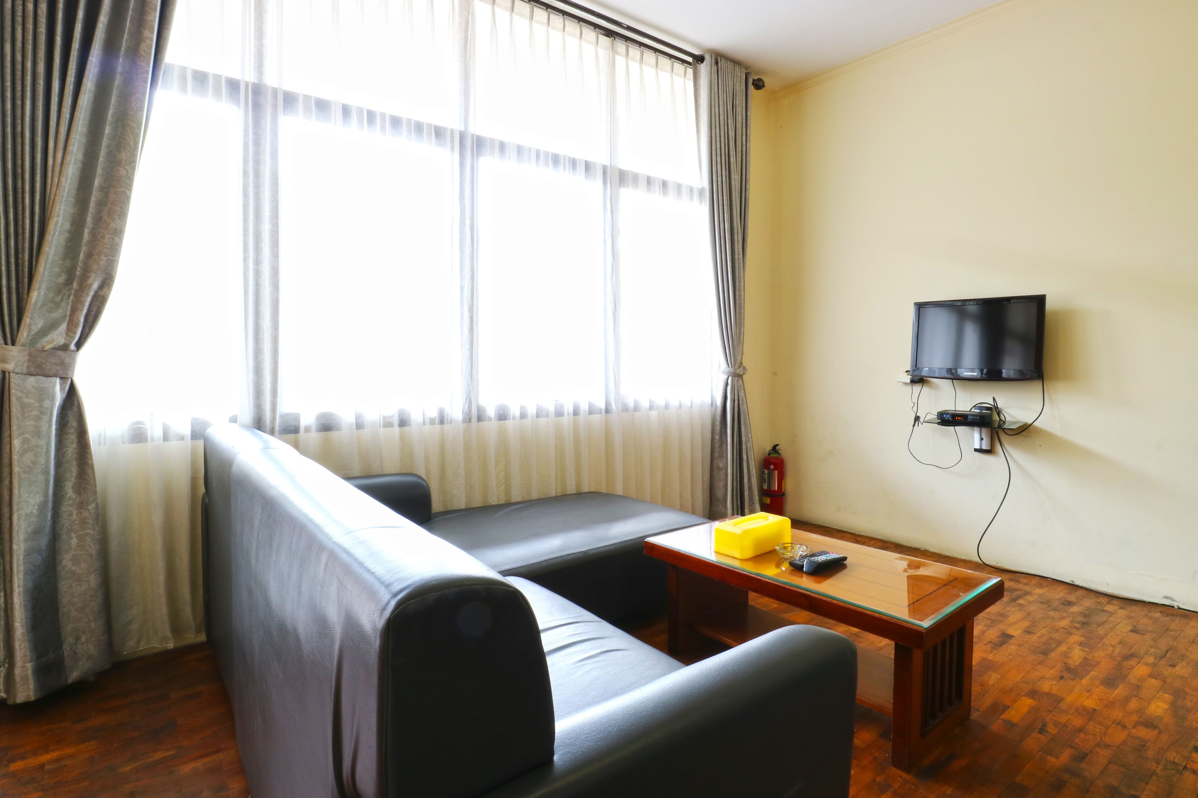 A room with a sofa, a small table, a television mounted on the wall, and a window with curtains.