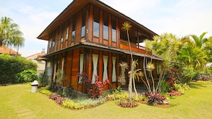 A two-story wooden house with a balcony, surrounded by a well-maintained garden with various plants and a lawn.