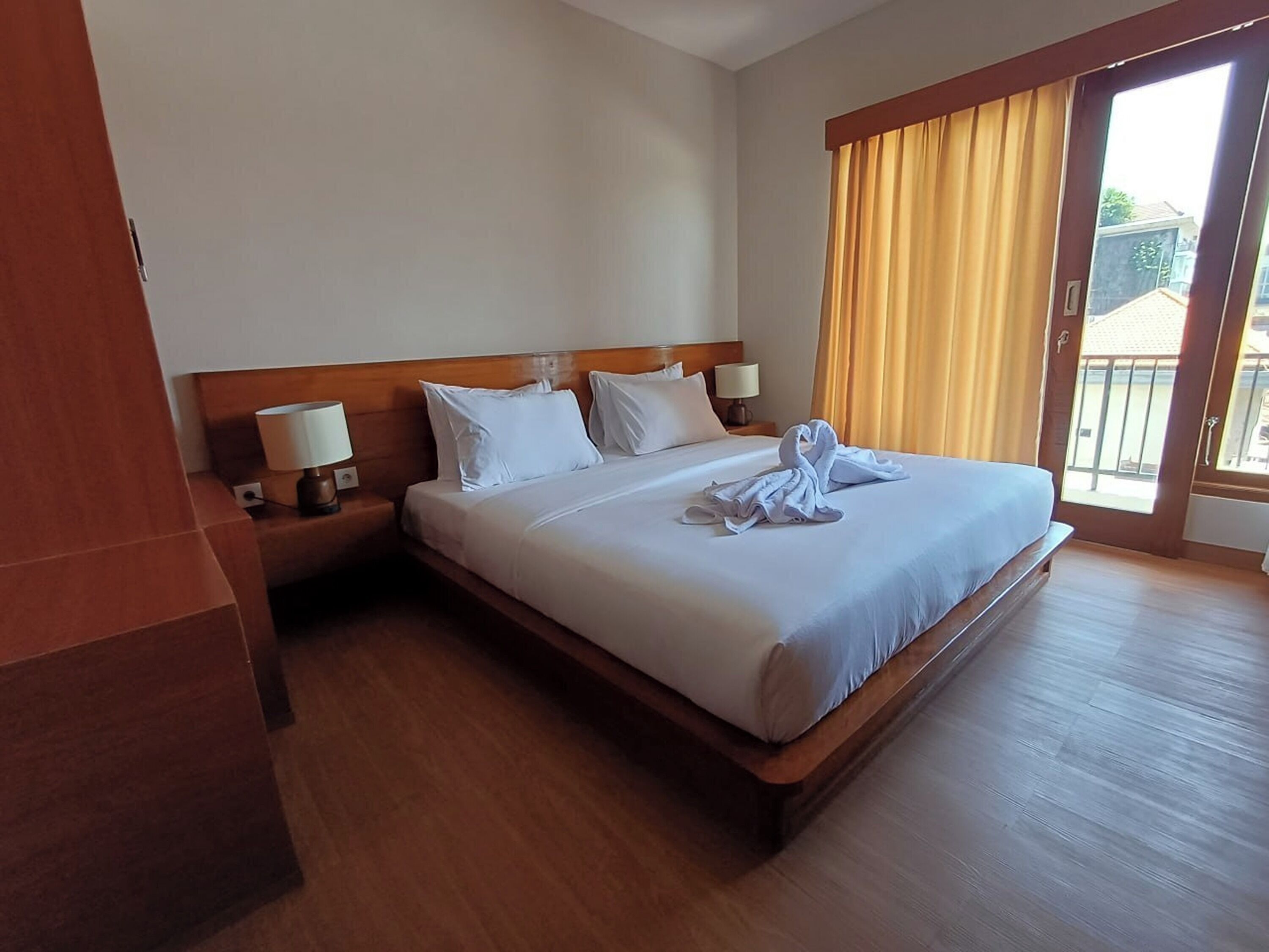 A hotel room with a large bed, two bedside lamps, a wooden headboard, and a view through a sliding glass door.