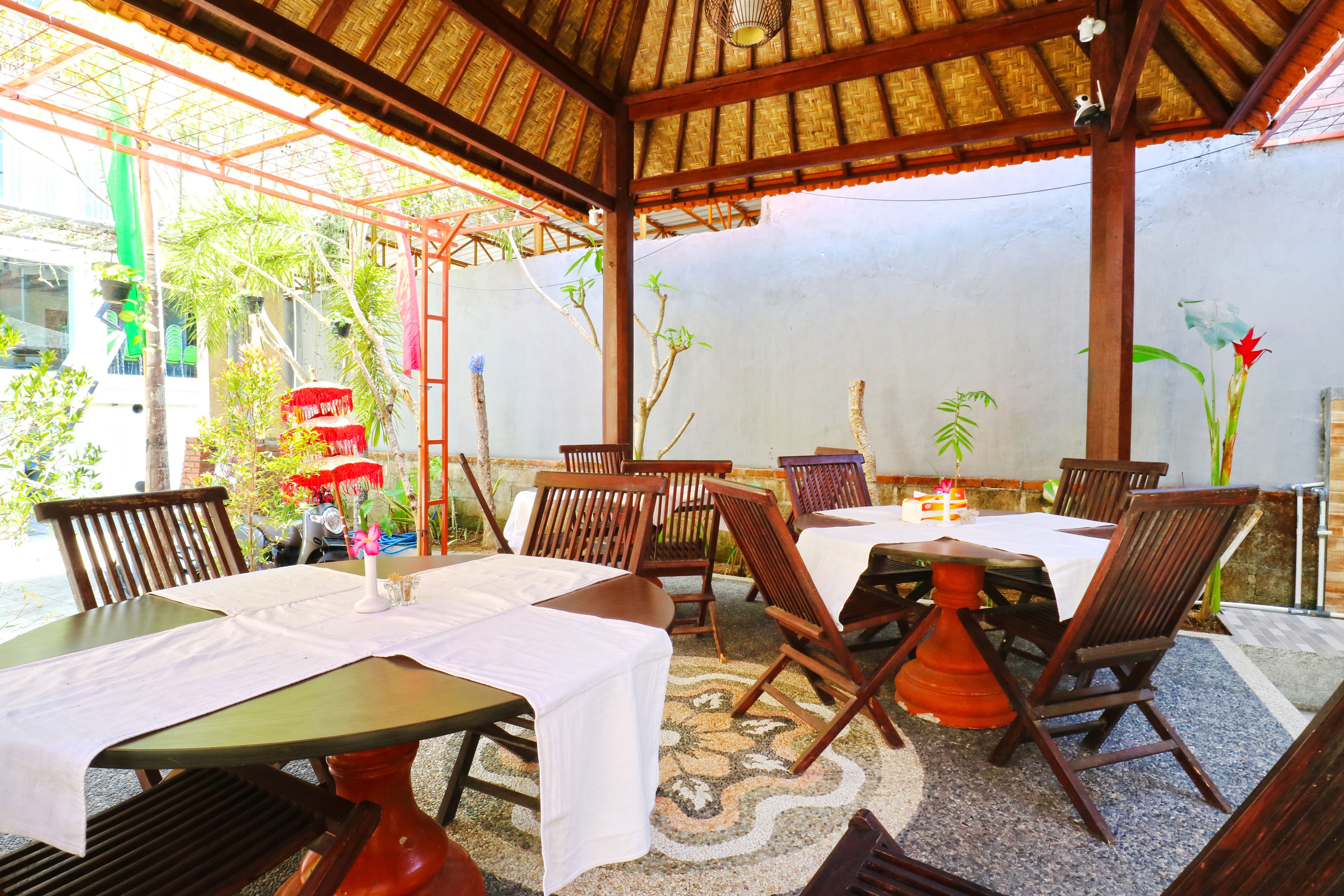 An outdoor dining area with wooden tables and chairs, covered by a thatched roof.
