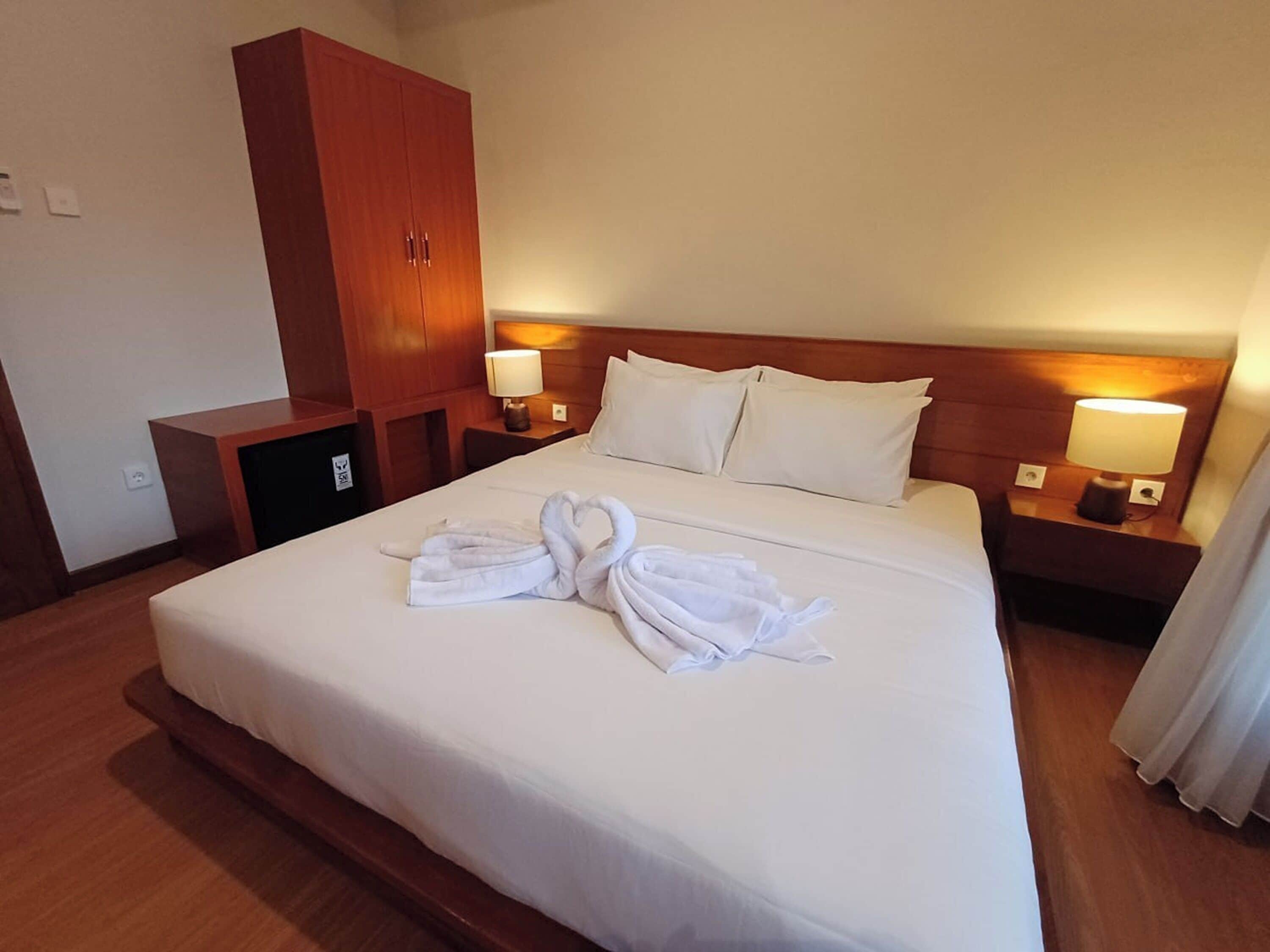 A hotel room with a large bed, two bedside tables with lamps, a wardrobe, and a pair of towels arranged on the bed.