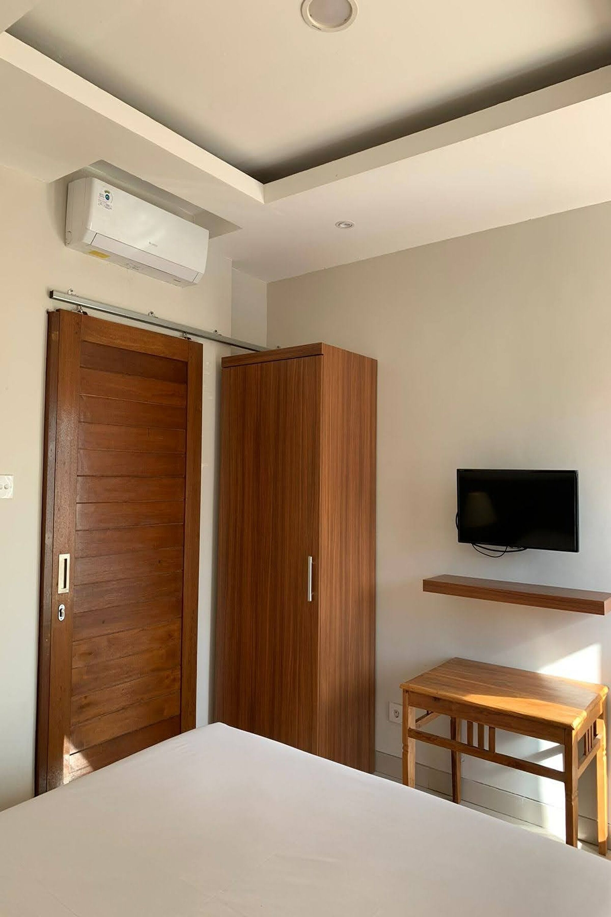 A bedroom with a bed, a wooden wardrobe, a television mounted on the wall, and a small wooden table.