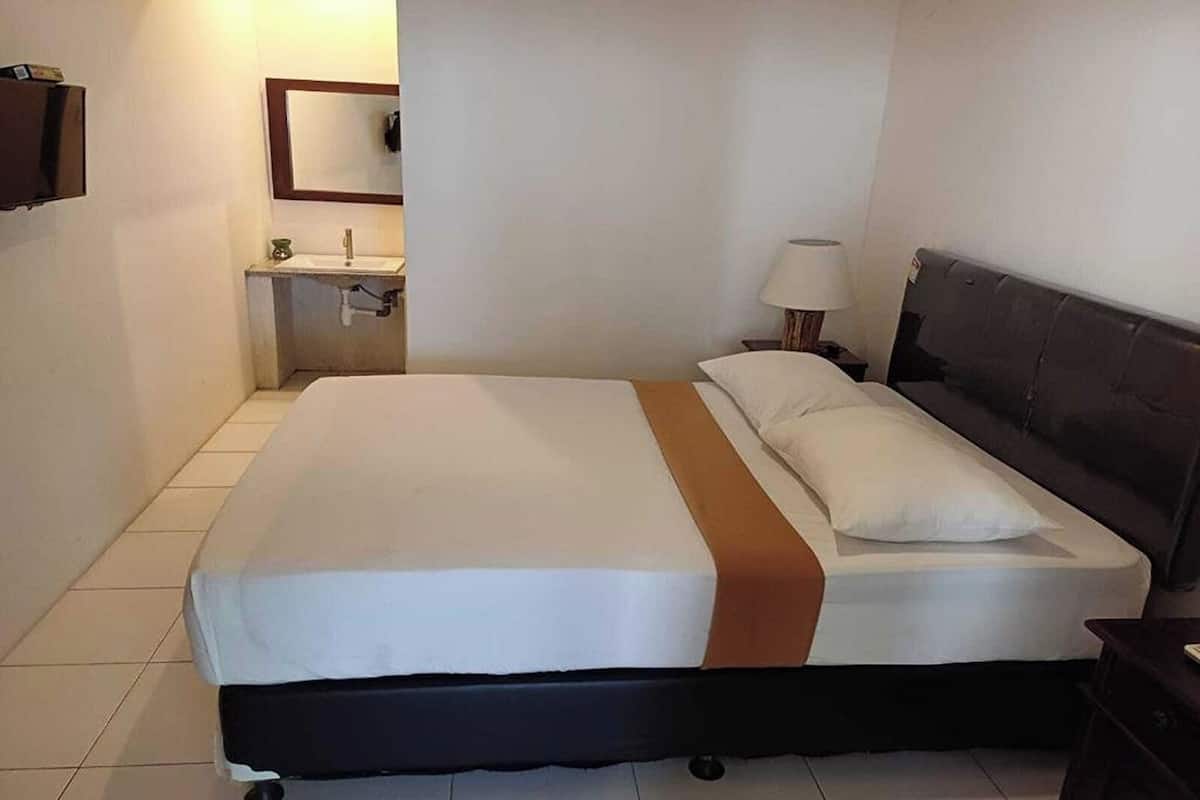 A hotel room with a bed, a nightstand, a lamp, a mirror, and a bathroom with a sink and toilet.