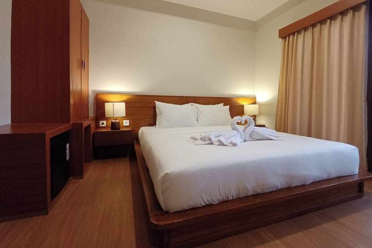 A hotel room with a large bed, wooden headboard, two bedside tables with lamps, a wardrobe, and a large window with curtains.