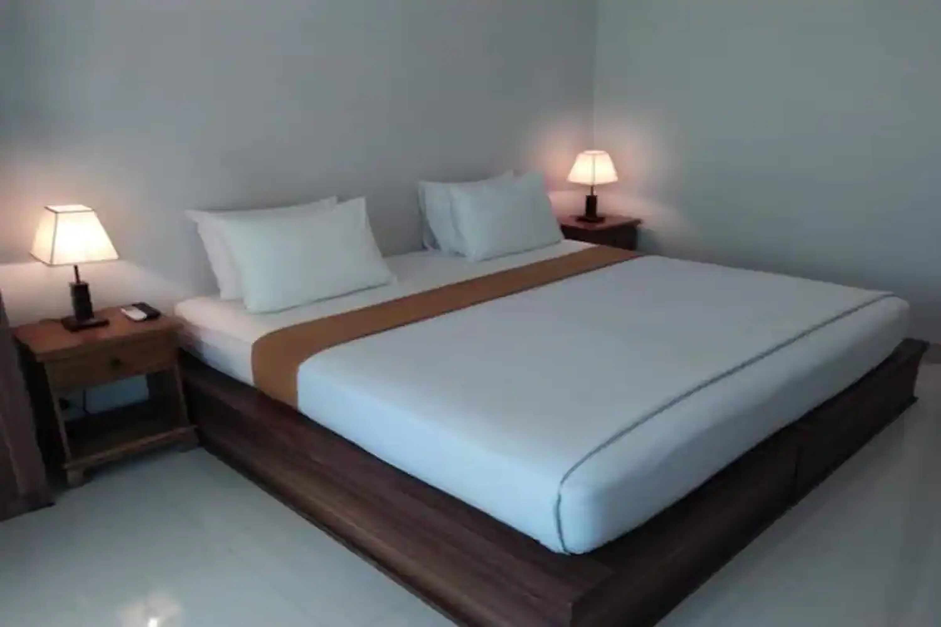 A neatly made bed with a wooden frame, two bedside tables with lamps, and a plain wall.