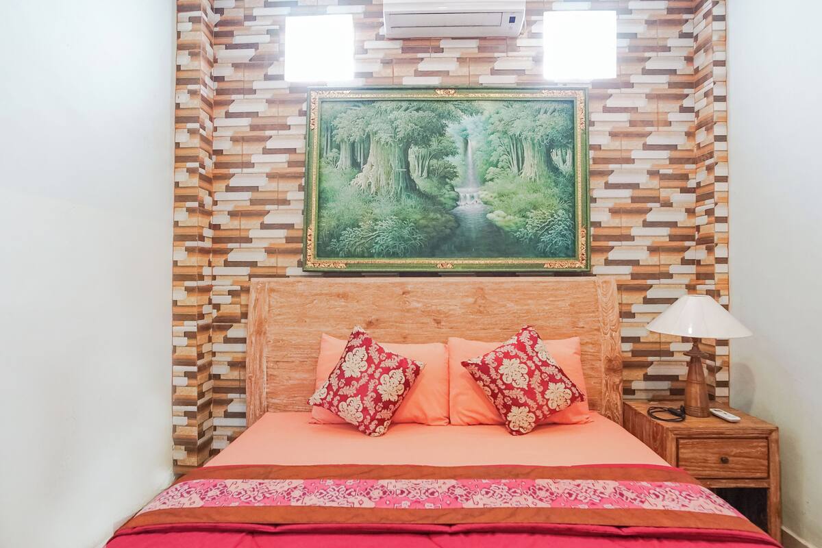 A bedroom with a bed, pillows, a nightstand, a lamp, and a wall-mounted painting of a forest scene.