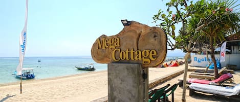 Un letrero de madera que dice "Mega Cottage" en una playa con un bote en el agua y sillones de descanso cerca.