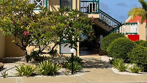 Une maison à deux étages, avec un toit recouvert de tuiles rouges, un balcon doté d’une balustrade blanche et un petit jardin où poussent différentes espèces de plantes.