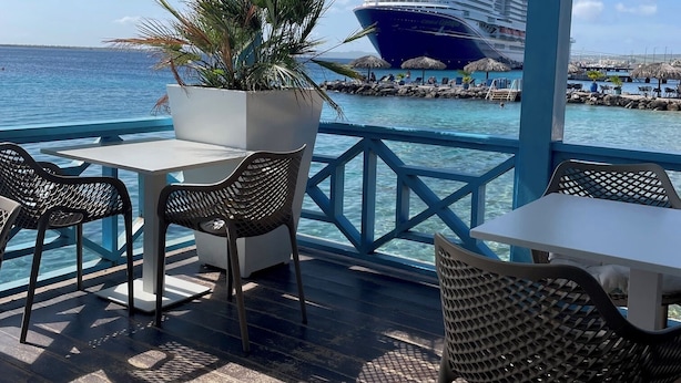 Un espace extérieur aménagé, abrité et doté de tables et de chaises, offrant une vue sur une étendue d’eau et un navire de croisière.