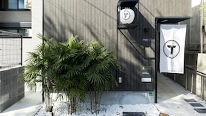 A modern building facade with a vertical wooden panel design, a metal gate, and a small garden area with white pebbles and green plants. - Trip Pod Minoshima A (Fukuoka)