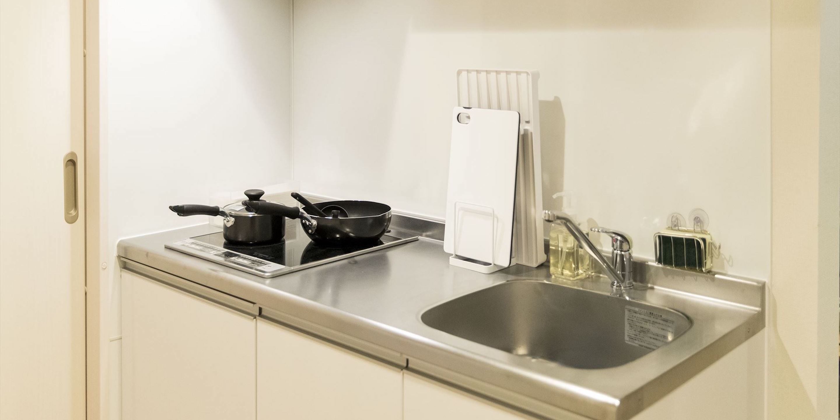 A compact kitchen with a stove, a frying pan, a kettle, a sink, and a white towel hanging on the wall.