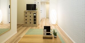 A hotel hallway with wooden flooring, a wooden cabinet with drawers, a television on top, a small plant, and a wall-mounted light fixture. - Trip Pod Minoshima A (Fukuoka)